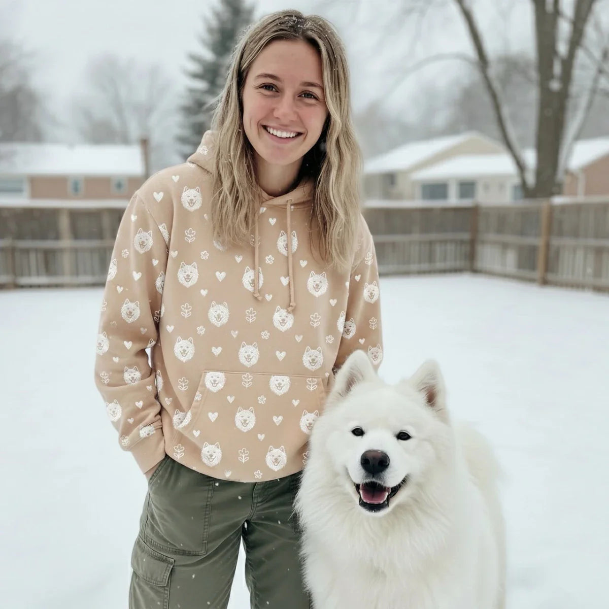 A smiling woman stands in the snow with her white Samoyed, wearing a tan hoodie with a repeat Samoyed face and heart pattern. This Samoyed Gift collection features breed-specific apparel, home decor, and accessories.