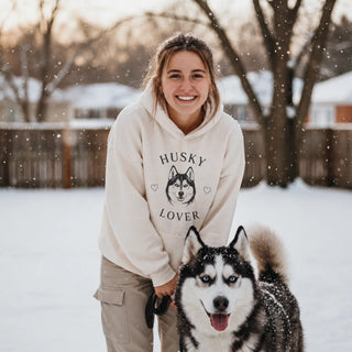 A smiling woman wearing a "Husky Lover" graphic hoodie stands in a snowy backyard with her Siberian Husky. This Husky Gift collection features breed-specific apparel, home decor, plush toys, and holiday ornaments.